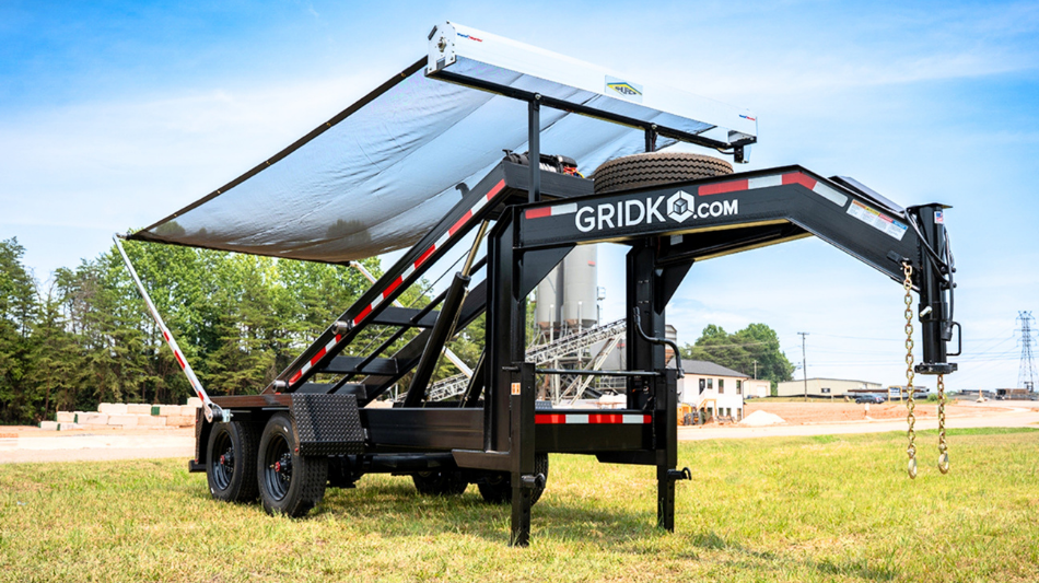 Black Gridko gooseneck roll-off trailer with its tarp cover system extended, parked on grass with an industrial facility and silos in the background.